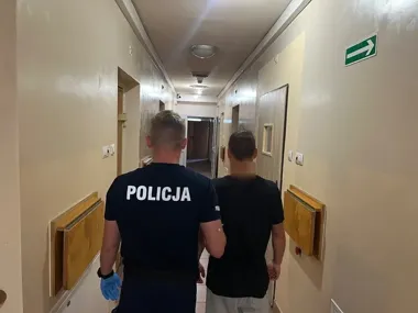 Policjant prowadzi mężczyznę z zamazaną twarzą korytarzem budynku.