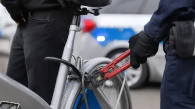 Ręka w czarnej rękawicy trzyma czerwone nożyce do metalu przy zabezpieczeniu roweru, w tle rozmyty radiowóz.