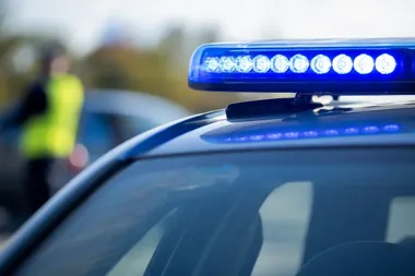 Zbliżenie na niebieski sygnalizator świetlny na dachu policyjnego radiowozu, w tle rozmyta postać funkcjonariusza w kamizelce odblaskowej.