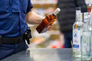 Funkcjonariusz w niebieskim mundurze trzyma w ręku butelkę z bursztynowym płynem przy ladzie z butelkami alkoholu.