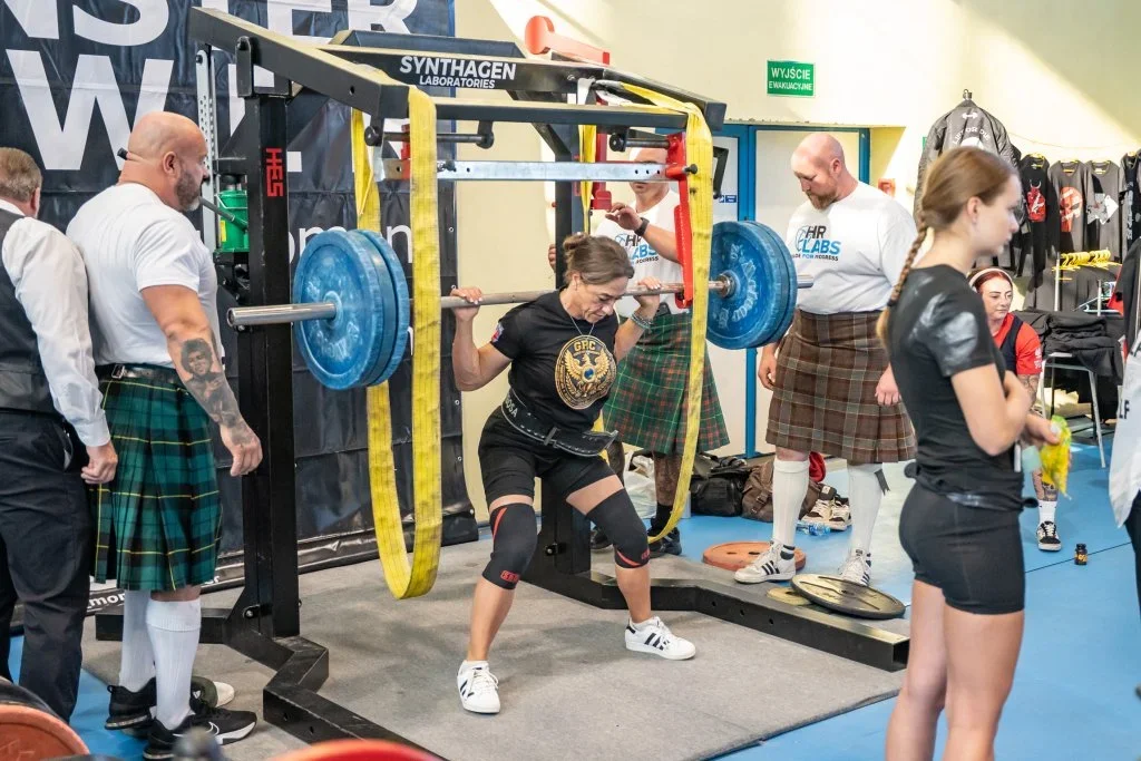 Kobieta w stroju sportowym wykonuje przysiad ze sztangą w siłowni, podczas gdy mężczyźni w kiltach obserwują ćwiczenie.